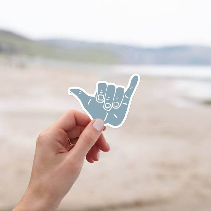 Person holding Blue Shaka Surf Sticker with beach background