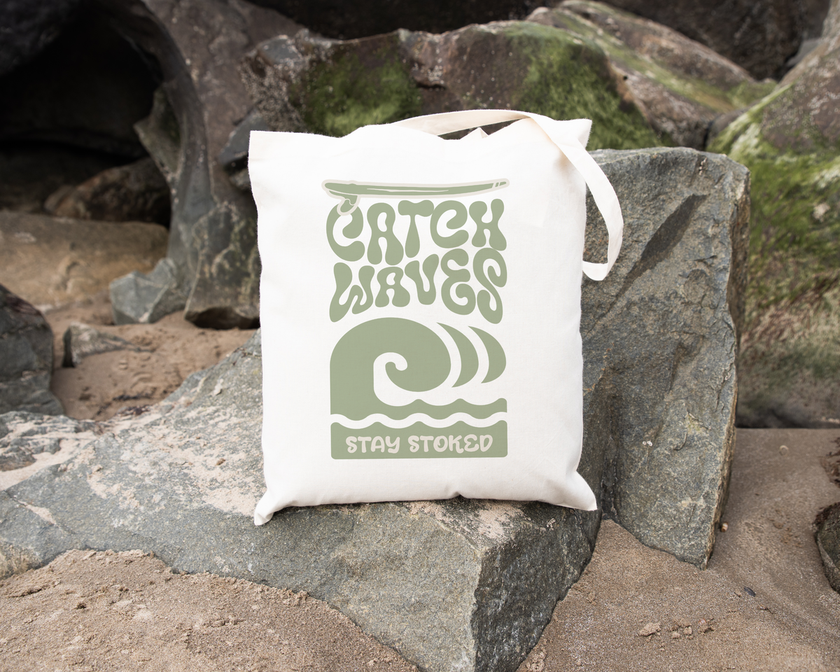 Catch Waves Stay Stoked canvas tote bag on rocks, featuring stylish wave design, perfect for beach outings, made of organic cotton.