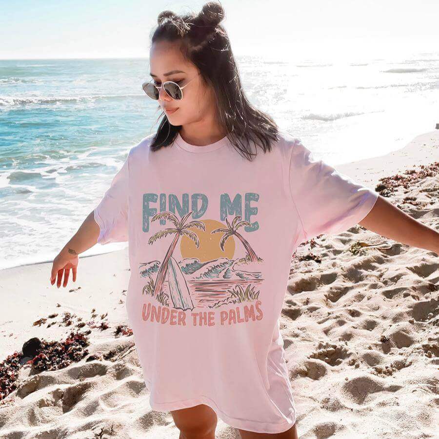 Woman wearing Under the Palms Soft Pink Graphic Tee on a beach, featuring a vibrant tropical design and palm trees.