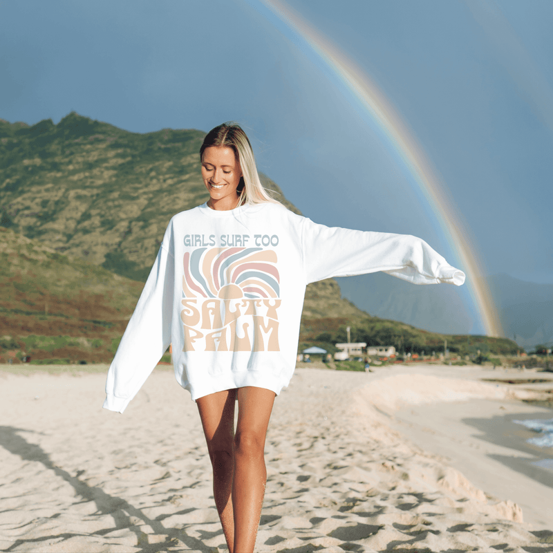 Young woman wearing a Girls Surf Too white crewneck sweatshirt on a beach with a rainbow background.