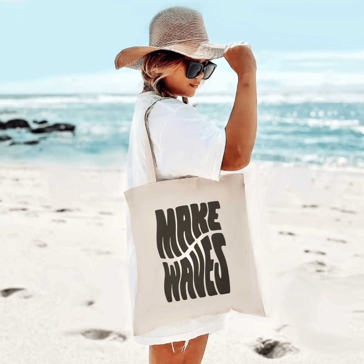 Woman at beach with Make Waves Canvas Tote Bag, white dress, straw hat, sunglasses, holding organic cotton tote with stylish design