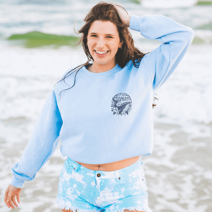 Woman wearing Protect the Oceans light blue crewneck sweatshirt at beach, supporting ocean conservation with casual style.