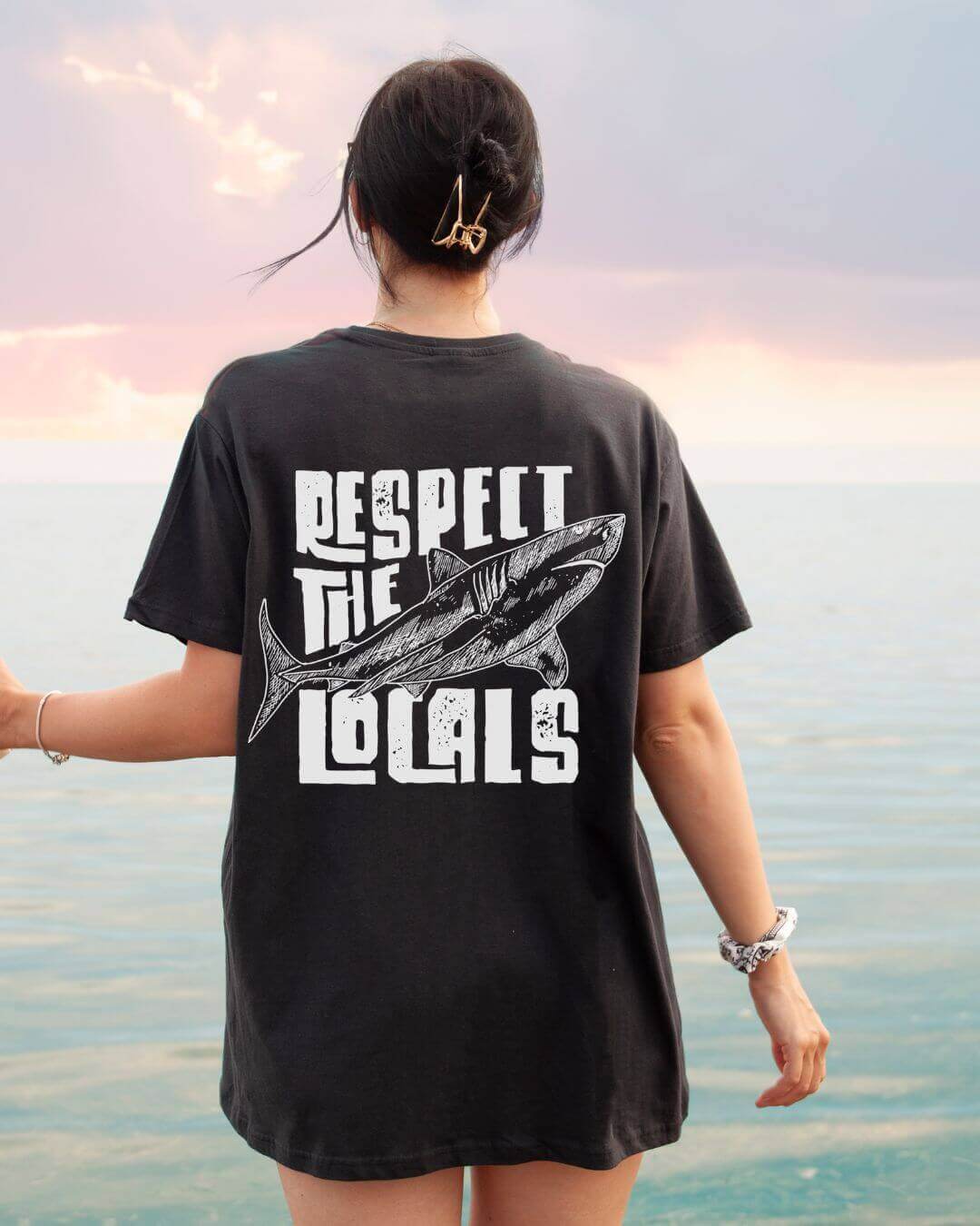 Woman wearing a vintage gray tee with "Respect the Locals" shark graphic, standing by the ocean under a sunset sky.