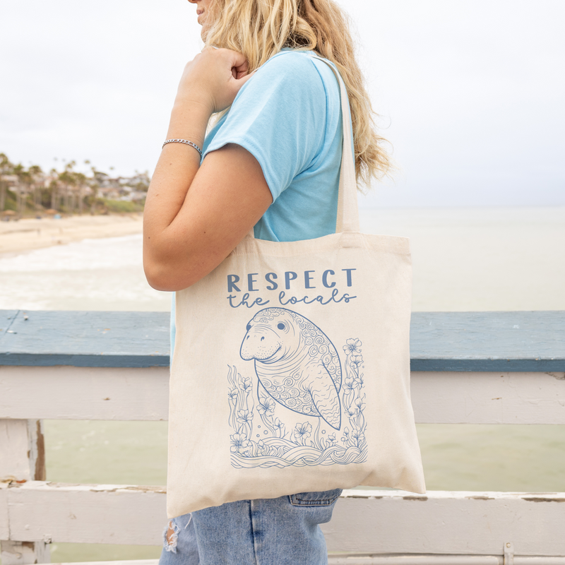 Woman holding Respect the Locals Manatee Canvas Tote Bag at the beach, eco-friendly and durable, perfect for everyday use.