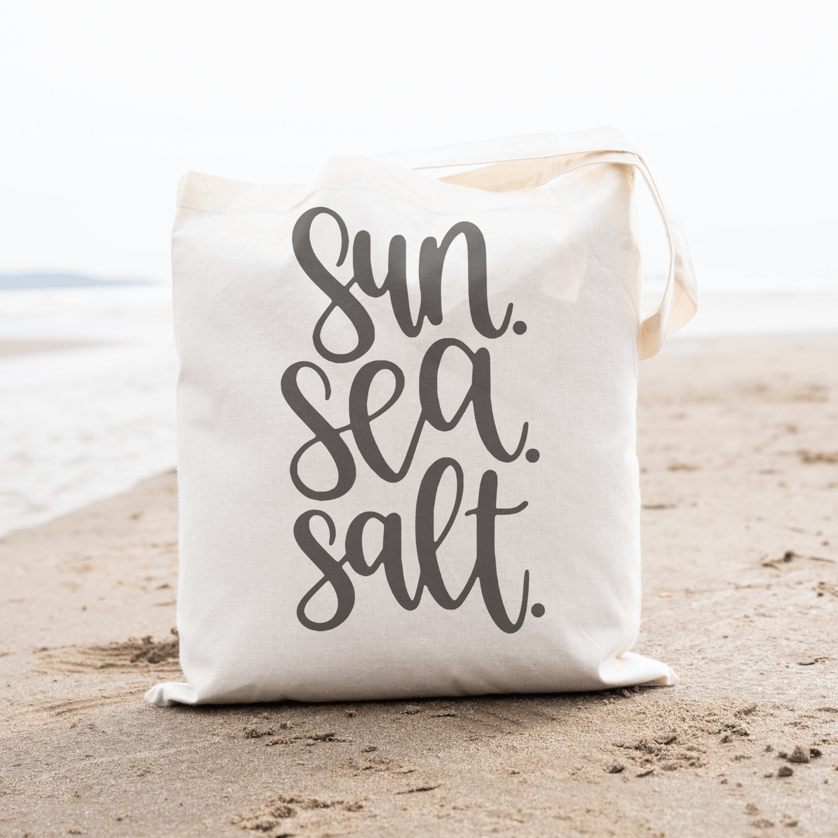 Sun Sea Salt canvas tote bag on sandy beach, perfect for beach days or errands, made from durable organic cotton with stylish design.