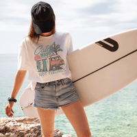 Woman in Catch Waves Not Feelings white graphic tee holding a surfboard, standing by the ocean.